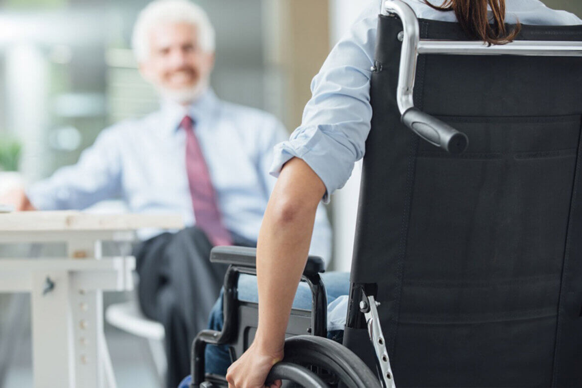 Persona su sedia a rotelle in contesto lavorativo insieme ad un collega che si vede sullo sfondo della foto